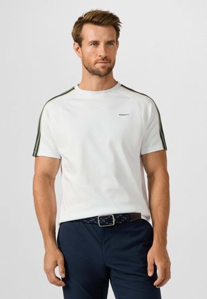 Man met kort bruin haar, met een wit kortemouwhemd met groene schouderstrepen en marineblauwe broek met gevlochten riem, staand tegen een effen achtergrond.