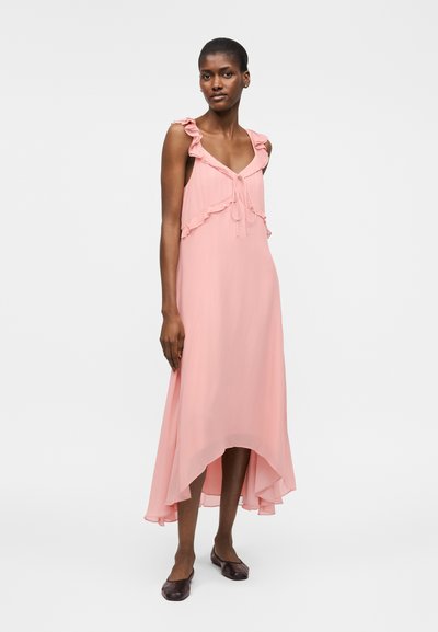 Γυναίκα που φορά αμάνικο ροζ φόρεμα με ασύμμετρο μήκος, λεπτομέρειες με βολάν και δετή λαιμόκοψη, συνδυασμένο με σκούρα επίπεδα παπούτσια, στέκεται σε λευκό φόντο.