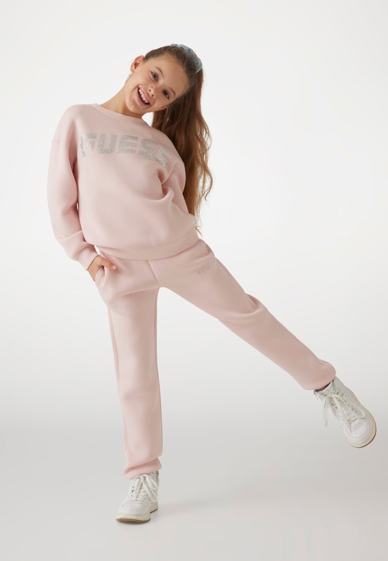 Rosa Trainingsanzug mit einem Sweatshirt im Crewneck-Stil, das mit silberner "GUESS"-Schrift versehen ist, und passenden tapered Pants, kombiniert mit weißen Sneakers.
