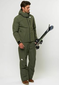 Olíva zöld sídzseki magas gallérral és cipzáras zsebekkel, hozzá illő nadrággal párosítva. Kiegészítők között szerepel fekete síléc és kötés.