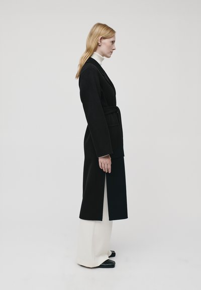 Black long coat with a belt, featuring a straight silhouette and split sides, worn over a white turtleneck and wide white pants.