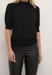 Sort kortærmet sweater med høj hals, lavet af struktureret stof, med puffede ærmer og en tætsiddende talje. Bæres med sorte leggings.