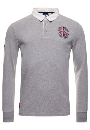 Homme portant un polo à manches longues gris clair, pantalon vert olive et baskets blanches, debout avec une main dans la poche contre un mur en bois.