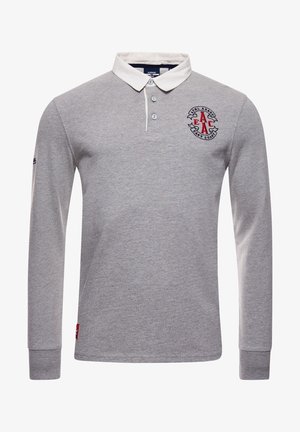 Homme portant un polo à manches longues gris clair, pantalon vert olive et baskets blanches, debout avec une main dans la poche contre un mur en bois.