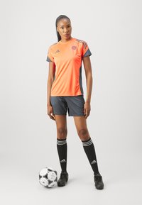 Πορτοκαλί και γκρι αθλητική φανέλα με κοντά μανίκια, συνδυασμένη με γκρι σορτσάκι, μαύρες κάλτσες με λευκές ρίγες και μαύρα παπούτσια ποδοσφαίρου, δίπλα σε μια μπάλα ποδοσφαίρου.