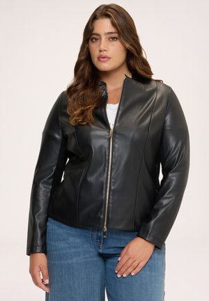 Frau mit langen braunen Haaren, die eine schwarze Lederjacke mit Reißverschluss über einem weißen Top und blauen Jeans trägt, steht vor einem einfarbigen Hintergrund.