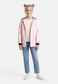 Νεαρή κοπέλα με πλεκτά μαλλιά, φορώντας ροζ μπουφάν με κουκούλα, λευκό πουκάμισο, μπλε τζιν, λευκές κάλτσες και λευκά αθλητικά παπούτσια, στέκεται μπροστά σε λευκό φόντο.