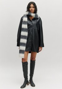 Schwarzes Lederhemdkleid mit Knopfverschluss, ausgestattet mit einer Vordertasche. Kombiniert mit einem gestreiften schwarz-weißen Strickschal. Knielange schwarze Stiefel.