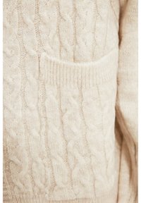 Svetr z pleteniny v měkké krémové barvě, který má přední kapsu a žebrované okraje. Texturovaný vzor přidává designu na rozmanitosti.