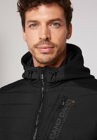 Musta pehmustettu takki korkealla kauluksella, vetoketjukiinnityksellä ja verkkoyksityiskohdilla. "CAMP DAVID" -logo rinnassa, jossa on tyylikäs muotoilu.