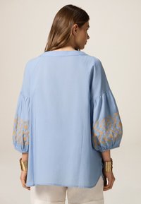 Blusa azzurro chiaro con maniche a sbuffo e ricami geometrici dorati sui polsini. Tessuto morbido, vestibilità rilassata e design con schiena scoperta.