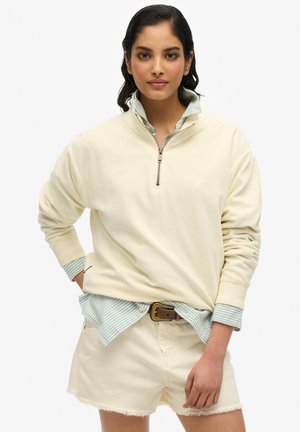Helles Creme Quarter-Zip Sweatshirt mit gerippten Bündchen, über einem türkis gestreiften Hemd mit Kragen getragen; kombiniert mit cremefarbenen Shorts.
