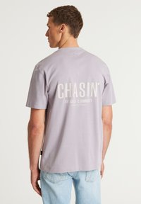 T-shirt di cotone lavanda con maniche corte. Grande scritta bianca "CHASIN'" sulla schiena, accompagnata da un testo più piccolo sottostante. Design casual.