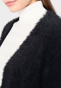 Kvinna som bär en vit ribbad turtleneck-tröja under en svart fluffig cardigan, med rakt brunt hår synligt på ena sidan.