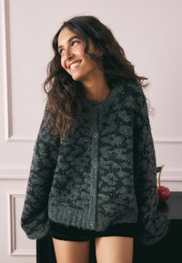 Pletena siva kardigan jakna s teksturiranim uzorkom, slobodnog kroja, velikim rukavima i kopčanjem sprijeda. Sadrži suptilne sjajnosti kroz cijelu jaknu.