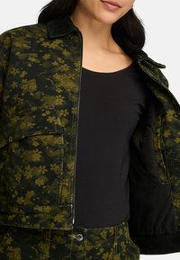 Giacca verde e nera con motivo floreale, dotata di zip frontale, colletto in velluto a coste e taschino sul petto. Camicia nera indossata sotto.