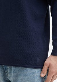 Navy textuurtrui gemaakt van een geweven stof, met een subtiel logo aan de onderkant en aansluitende lange mouwen. In combinatie met lichte denim.