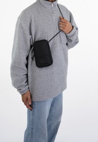 Svart crossbody-väska med en slät yta, prydd med ett logotyget, bärs över en grå fleece-tröja, tillsammans med ljusblå jeans.