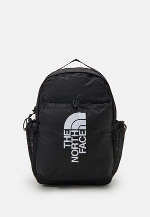 Sac à dos noir avec poche avant zippée, poches latérales en mesh, poignée supérieure et grand logo blanc "The North Face" à l'avant.