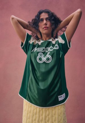 Žena s kučeravými vlasmi v zelenej futbalovej šatke "Mexico 86" a žltej pletenej sukni pózuje s rukami za hlavou na ružovom pozadí.