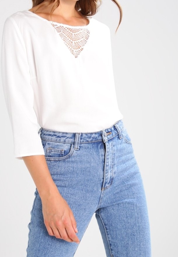 Femme portant un haut blanc à manches longues avec un détail en dentelle en V et un jean bleu clair taille haute, debout avec une main détendue.