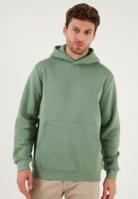 Vihreä huppari, jossa etutasku, valmistettu pehmeästä kankaasta. Varustettu kiristysnauhalla varustetulla hupulla sekä resorikäänteillä hihansuissa ja helmassa. Sileä pinta, rento muotoilu.