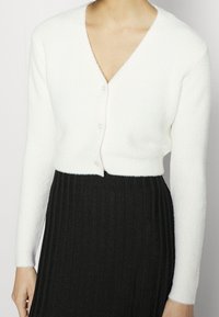 Eine Person trägt eine weiße, knopfförmige, strukturierte Strickjacke und einen schwarz, hochtaillierten, plissierten Rock, gezeigt von Hals bis Taille.