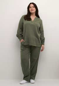 Olivgrünes, strukturiertes Zweiteiler-Outfit mit einem langärmeligen Oberteil mit V-Ausschnitt und geraden Hosen. Weiße Sneaker runden den Look ab.