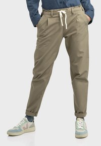 Beige, taillierte Hose mit einem Kordelzugbund, Falten und Seitentaschen. Getragen mit mehrfarbigen Turnschuhen und einem blauen Hemd.