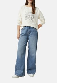 Frau, die lockere, hellblaue, weite Jeans, weiße Sneakers und ein langärmliges, weißes Oberteil mit einem New Yorker Skyline-Druck trägt, steht mit einer Hand auf der Hüfte.