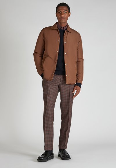 Bruine poff jacket met een kraag, een marineblauwe trui eronder en een broek met een bruin ruitpatroon, gecombineerd met zwarte nette schoenen.