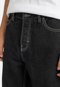 Jeans de mezclilla negra con costuras blancas y cierre de botón. Presenta cinco bolsillos y un corte relajado, resaltando un acabado de tela texturizada.