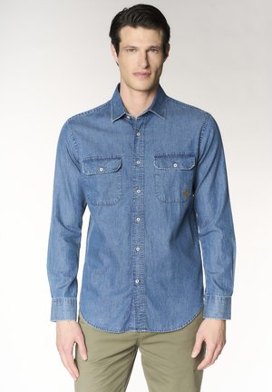 Uomo che indossa una camicia di jeans blu abbottonata con due tasche sul petto e pantaloni beige, in piedi davanti a uno sfondo semplice.