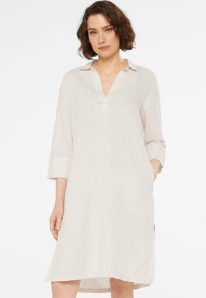 Woman with short brown hair wearing a loose, off-white, long-sleeve knee-length dress with a V-neck collar.