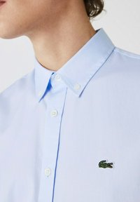 Lichtblauwe katoenen shirt met een button-down kraag, witte knopen en een klein groen geborduurd logo op de borst.