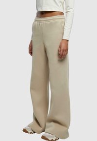Béžové široké tepláky s elastickým pásom, ktoré majú jemnú textúru a bočné vrecká, kombinované s béžovými a off-white teniskami.