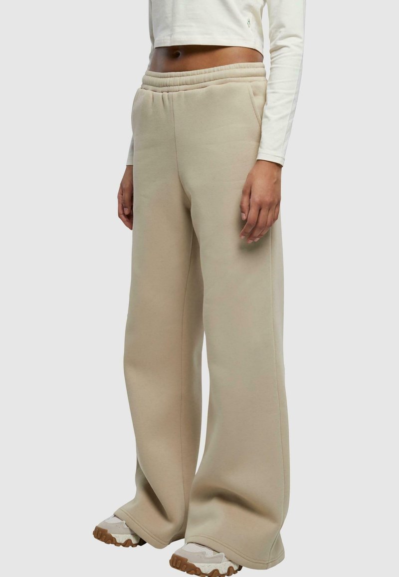 Béžové široké tepláky s elastickým pásom, ktoré majú jemnú textúru a bočné vrecká, kombinované s béžovými a off-white teniskami.