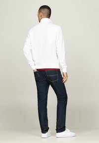 Sudadera blanca con cuello alto, combinada con jeans azul oscuro y zapatillas blancas. El conjunto se ve desde la parte de atrás.