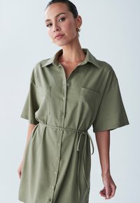 Olive roheline särgkleid, millel on lühikesed varrukad, nööbiga eesmine osa, kaks rinna taskut ja vööga talje, et luua määratletud kuju.