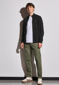 Černá zipová bunda přes bílé tričko, spárovaná s olivově zelenými kalhotami a béžovými teniskami. Minimalistický design bez viditelných vzorů.