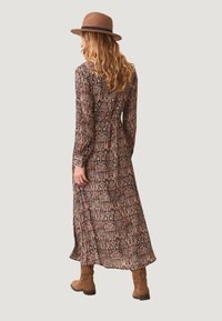 Vestido maxi de tejido oscuro con estampados florales y geométricos en rosa. Mangas largas, cintura ajustada y falda fluida. Combinado con un sombrero marrón y botas.