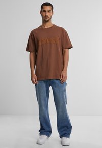 Ruda medvilninė trumpomis rankovėmis marškinėliai su reljefiniu logotipu panašios spalvos, derinami su šviesiai mėlynomis džinsinėmis kargo kelnėmis ir baltais sportbačiais.