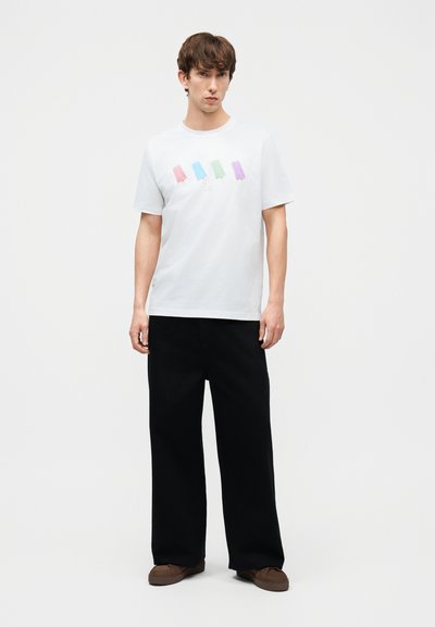 Νεαρός άνδρας στέκεται φορώντας λευκή μπλούζα με τέσσερις πολύχρωμες εικονογραφήσεις παγωτών σε ραβδάκι, μαύρο παντελόνι με φαρδιά πόδια και καφέ παπούτσια, μπροστά σε απλό φόντο.