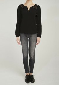 Svart långärmad topp med spetsdetaljer och V-hals, i kombination med åtsittande grå jeans och svarta skor, stående mot en neutral bakgrund.