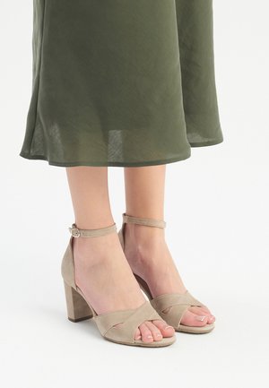 Sandali con tacco in suede beige, con punta aperta, cinture incrociate, fibbia alla caviglia e tacco a blocco. Abbinati a una gonna verde oliva.