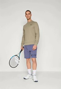 Grønn langermet sweatshirt, blå shorts med "BORG" trykk, hvite treningssko, holder en tennisracket. Enkel design og tettsittende form.