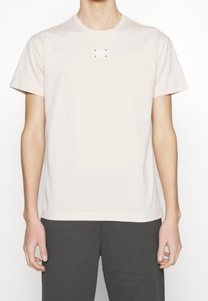 Light pink cotton t-shirt with short sleeves, round neckline, and small rectangular tag with metallic accents on the chest.