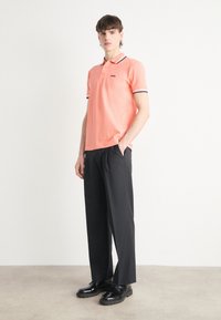 Helles rosa Polohemd mit schwarzen Akzenten, kombiniert mit lockeren schwarzen Hosen und glänzenden schwarzen Schuhen. Das Hemd hat ein kleines Logo.