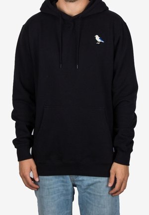 Mand iført sort hættetrøje med frontlomme og lille hvid og blå mågelogo på brystet, kombineret med lyseblå jeans.