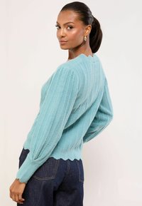 Hellblauer gerippter Pullover mit gewelltem Saum und langen Ärmeln, aus weichem Material. Trägt man über dunkelblauer Jeans vor neutralem Hintergrund.